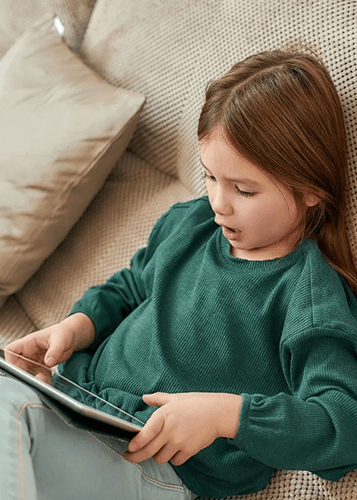 Kislány mélyen elmerülve nézi a tabletjét, miközben a kanapén ül. Digitális oktatás, online tanulás, gyerekek és technológia, otthoni kikapcsolódás.