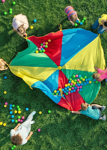 Gyerekek játszanak a fűben egy színes ejtőernyővel és sok színes labdával, felülnézetből. Vidám szabadtéri program gyerekeknek, nyári tábor.