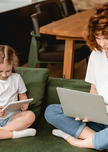 Egy anya és gyermeke ülnek egy zöld kanapén, mindketten digitális eszközöket használnak. A nő egy laptopot tart az ölében, a gyermek pedig egy tabletet. A háttérben egy étkezőasztal és székek láthatók.